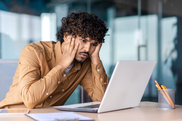 Confused muslim employee resting head on hands while looking at pc screen with incomprehension indoors. Curly male trainee desperately trying understanding project details on first working day. - obrazy, fototapety, plakaty