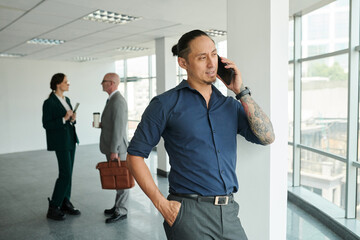 Young Asian male solopreneur in shirt and pants speaking on mobile phone while standing in front of camera in office center
