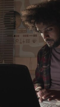 Vertical medium shot of Middle Eastern programmer with messy curly hair sitting in dark room with warm yellow light from bedstand lamp typing on old laptop