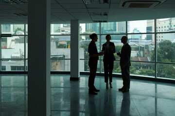 Outlines of three colleagues standing in spacious office center against large windows and discussing working points at meeting