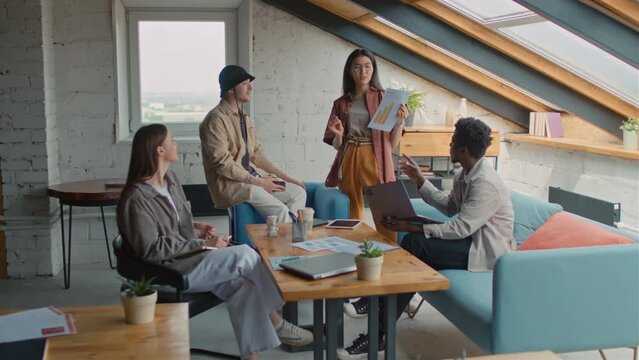 Full zoom shot of diverse startup team having business meeting in loft coworking office - Asian woman presenting graph with growth strategy, others listening, asking questions and arguing
