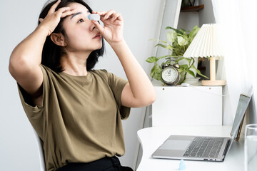 Asian woman applying eyedrops for her eye while working on laptop screen to relieve dry eyes and eyestrain