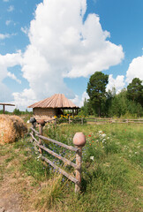 Village yard with buildings. Clay pots hang on the fence. Yellow hay. Wildflowers in a green meadow. Blue sky with white clouds. There is a forest in the background.