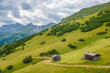 Fototapeta na wymiar Picturesque summer mountain landscape from Stubai Alps, Tyrol, Austria. A green mountainside, two small wooden houses and a road winding past them, scenic alpine peak and cloudy sky in background.