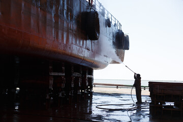 Worker cleans vessel hull in dry dock with high-pressure washer. Ship maintenance, maritime...