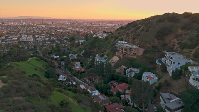 Aerial panorama of residential homes above city of Los Angeles cityscape shot from lush hiking trail in Hollywood Hills at sunset with camera flying backward.