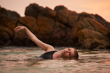 Woman in black swimsuit enjoys serene sea dip at sunset. Ocean bathing scene reflects wellness,...