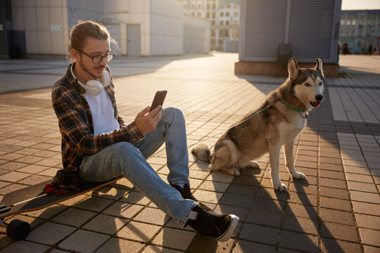 Cheerful young hipster guy using smartphone while resting after skateboarding