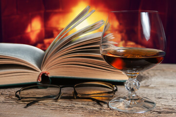 Glass of brandy, eyeglasses and old books in front of burning fireplace. Relaxed cozy atmosphere concept