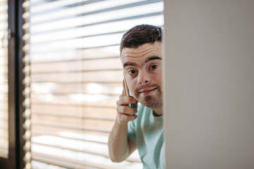Young man with Down syndrome sitting by window, making phone call with smartphone.