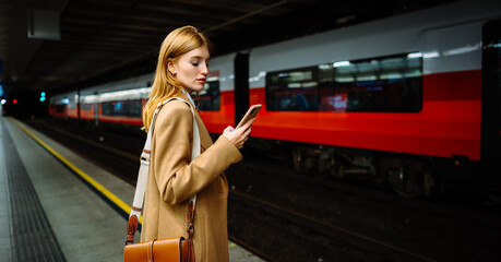 Female standing at subway platform with mobile phone, using social media and waiting train