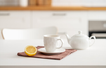 Fototapeta na wymiar A cup of tea with lemon and a teapot on a white table. Kitchen with wooden empty countertop and brick wall in the background.