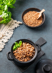 Buckwheat porridge in a black pan on a dark  background with raw grains and herbs.