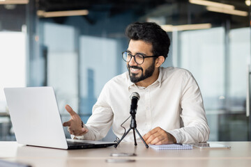 Obraz premium A smiling Indian young man is sitting in the office at a desk and talking through a microphone on a video call on a laptop. Conducts business training, holds a conference, online meeting