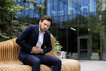 Grimacing and bent over, a young businessman in a suit sits on a bench on the street near an office...