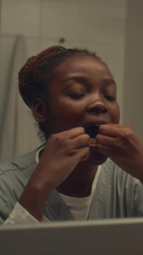 Closeup vertical shot of mirror reflection of young African American doctor or intern in medical uniform, with braids in bun, standing in bathroom and flossing teeth, before going to workCloseup verti