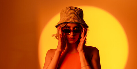 Fashionable model wearing trendy sunglasses and stylish hat against orange background in studio