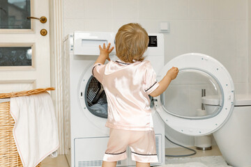 Rear view child girl little helper in pink pajama in laundry room near washing machine and dirty clothes in washing basket. washing and cleaning concept. daily routine. Copy space