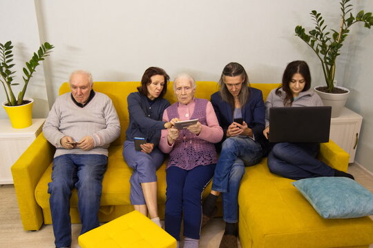Family members use various electronic devices. Grandparents, middle-aged woman and a young couple are sitting on the sofa in the room. An adult daughter helps her elderly mother use a tablet pc. - Powered by Adobe