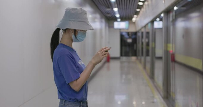Asian woman at metro station using a phone with surgical mask face protection.