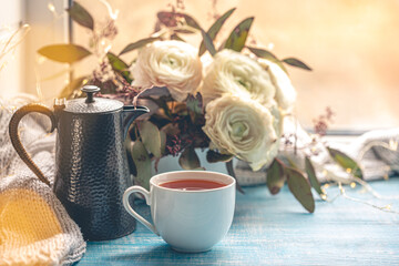 A cup of tea, a kettle and flowers by the window.