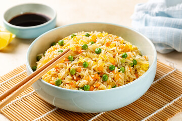Bowl with Fried rice with egg and vegetables, onion, bell pepper, carrot, green peas, soy sauce on background. Prepared and served in a light blue plate.