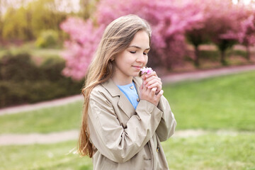 Outdoor fashion portrait little girl holding sakura flowers in his hands in blossom spring park. Young smile girl in a beige trench coat smiles and dreams standing at blossoming sakura garden. 