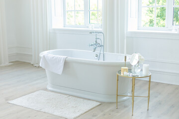 Stylish white bathtub against large windows and table with decoration. Concept of classic bath in modern apartment. Home decor ideas in contemporary bathroom scandinavian design. Template. Spa