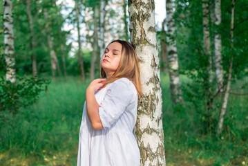 In summer, a young overweight girl stands by a tree in a white dress in the park. Space perspective. Ecology.
