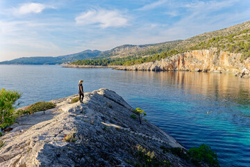 A woman wearing a hat admires the beach Malo Zarace in the Adriatic coast on Hvar island, Croatia.  Travel and summer holidays concept. - obrazy, fototapety, plakaty