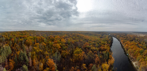 Autumn aerial panorama of river with colorful forest and grey cloudscape. Flying above autumnal vibrant rural Ukraine