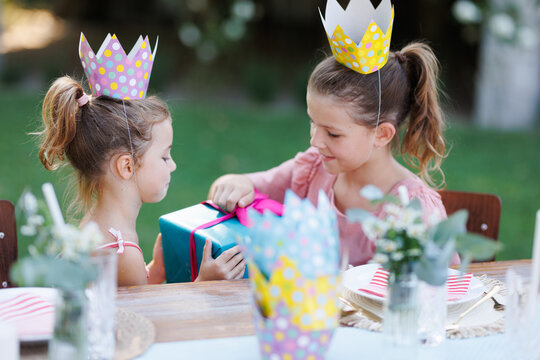 Little friends at birthday party outdoors at the garden. Cute girls with paper crowns opening birthday gift. Birthday garden party for children.