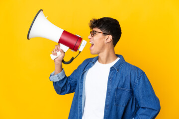 Young man over isolated yellow background shouting through a megaphone to announce something in lateral position