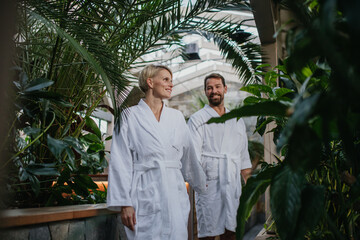 Beautiful couple in spa robes standing in hotel greenhouse, looking at each another, enjoying...