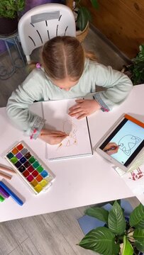 Online homeschooling, childhood hobby. Vertical of lovely little girl watches a video tutorial using a digital tablet to learn drawing with pencils indoors. Creativity, developing artistic skills