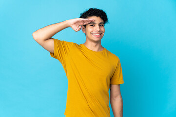 Young Venezuelan man isolated on blue background saluting with hand with happy expression