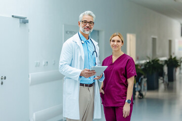 Portrait of medical team, doctor and nurse in uniform, standing in the hospital corridor, looking at the camera and smiling.