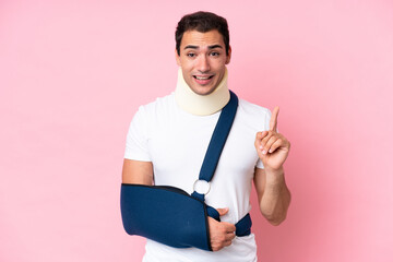 Young caucasian man with broken arm and wearing a sling isolated on pink background intending to...