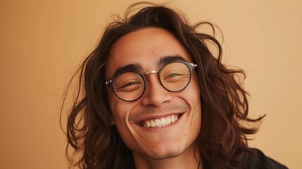 A young man with long curly hair wearing glasses smiling brightly against a warm blurred background.