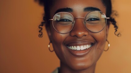 A joyful woman with curly hair wearing glasses and gold earrings smiling brightly against a warm orange background.