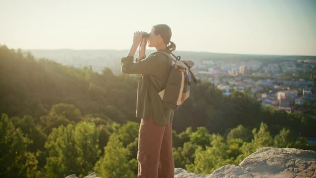 Beautiful Caucasian woman looking into binocular while standing on top of mountain. Satisfied young woman wearing backpack for her travel. Recreation on nature. Enjoying beautiful view.