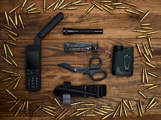 Close-up on a wooden background. Satellite phone with a large antenna, multi-tool, flashlight, atraumatic scissors, laser rangefinder, and hemostatic tourniquet. Frame of cartridges. - obrazy, fototapety, plakaty