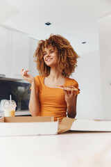 Attractive woman with curly hair enjoying a slice of pizza in her hand while wearing an orange shirt