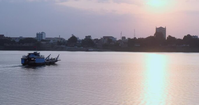 The local ferry, packed with passengers, is crossing the Mekong River. To the right of the river, the sun's rays paint the sky with the colors of sunset.