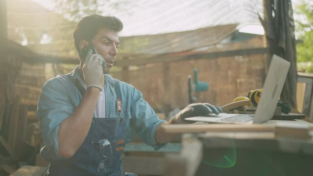 Young carpenter talking on smartphone and using laptop while working on woodworking in the furniture factory. Carpenter and woodcraft concept