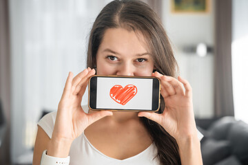 Young smiling woman holding mobile phone with valentine red heart on screen of smart phone. St. Valentine day concept