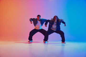 Energetic dance pair in denim jackets in squatting dance pose in neon-lit studio against gradient blue-orange background. Concept of youth culture, music, lifestyle, style and fashion, action.