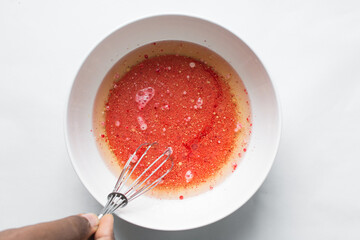 overhead view of oil, buttermilk and red food color being mixed in a white mixing bowl, process of making red velvet cake