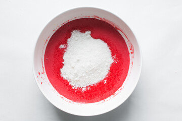 Top view of sugar being folded into Red velvet cake batter in white ceramic mixing bowl, process of making red velvet cake