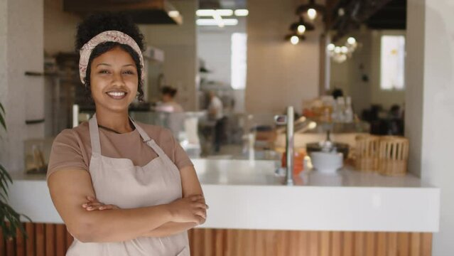 Portrait of young joyous woman in apron standing with arms crossed and smiling on camera while working in bakery